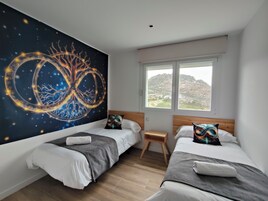 Un dormitorio con dos camas, un mural grande en la pared, una ventana con vistas a un paisaje montañoso y un taburete de madera pequeño.