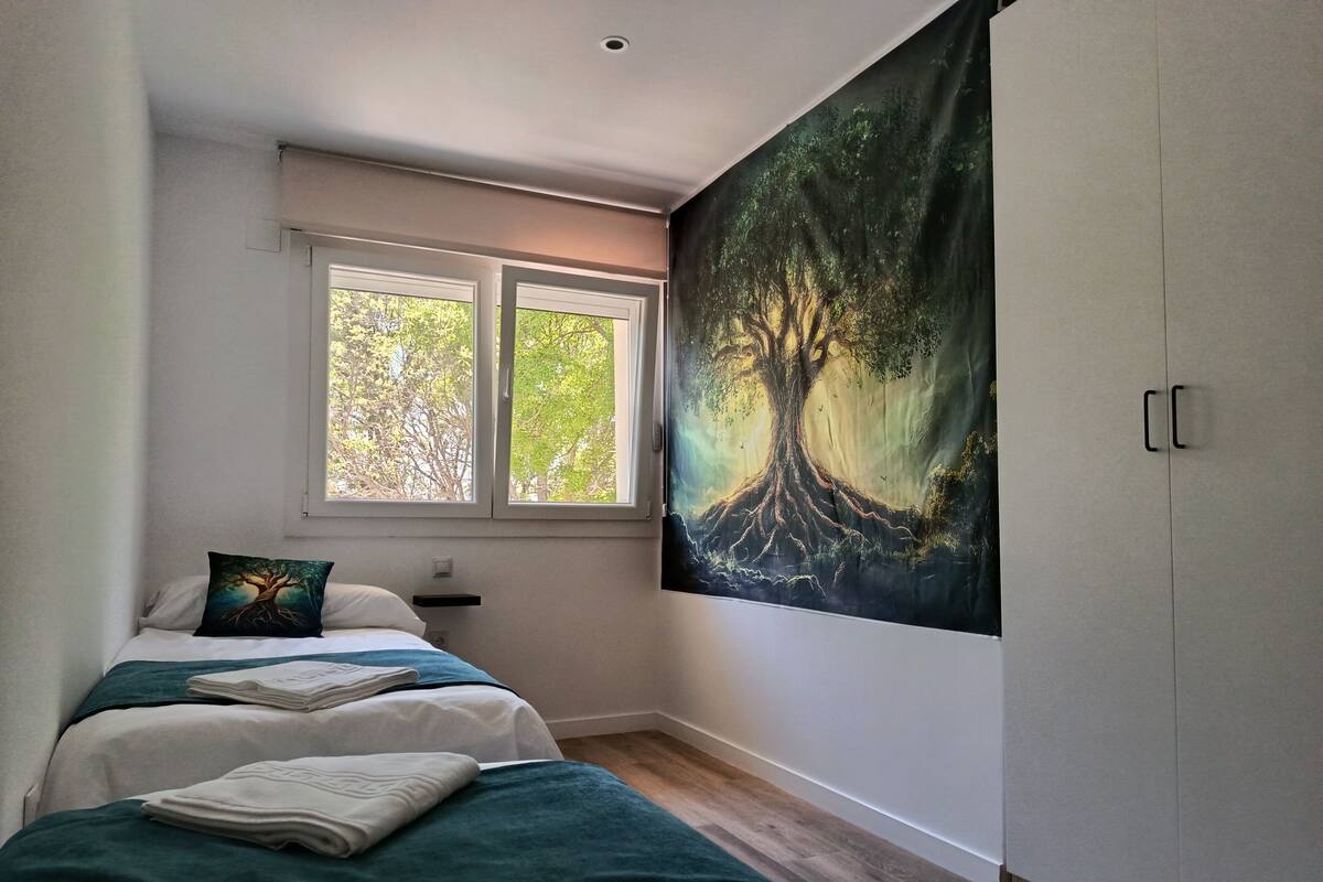 A bedroom with two beds, a window, and a large forest-themed tapestry on the wall.