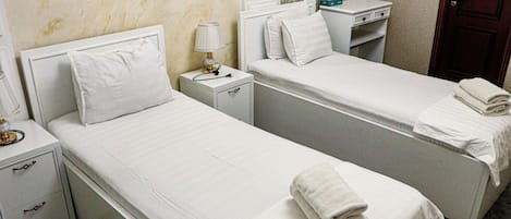 Habitación de hotel con dos camas individuales, una mesita de noche con lámpara, un escritorio pequeño y un armario.