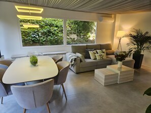 Ein modernes Wohnzimmer mit Esstisch, Sofa und Couchtisch, großen Fenstern mit Blick auf Grünflächen und einer Decke mit Einbauleuchten.