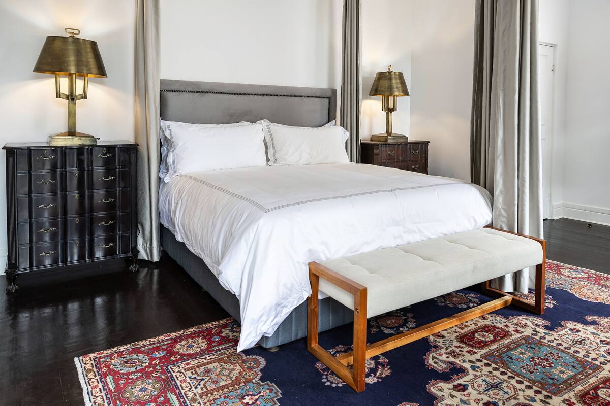 A neatly made bed with white linens, a bench at the foot, and a patterned rug on the floor.