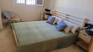 Una cama individual con cubrecama verde, dos mesitas de noche, una silla, un radiador y una ventana.