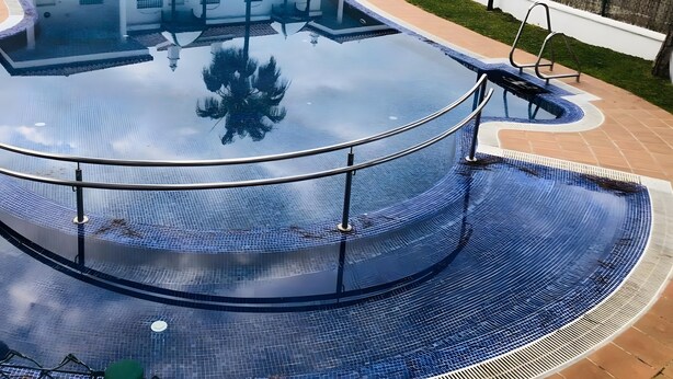 Una piscina con fondo de cristal, rodeada por un edificio blanco con balcones y un sendero pavimentado.