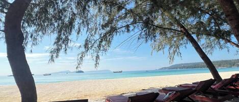 A beach with wooden lounge chairs under trees, a sandy shore, and boats in the water.