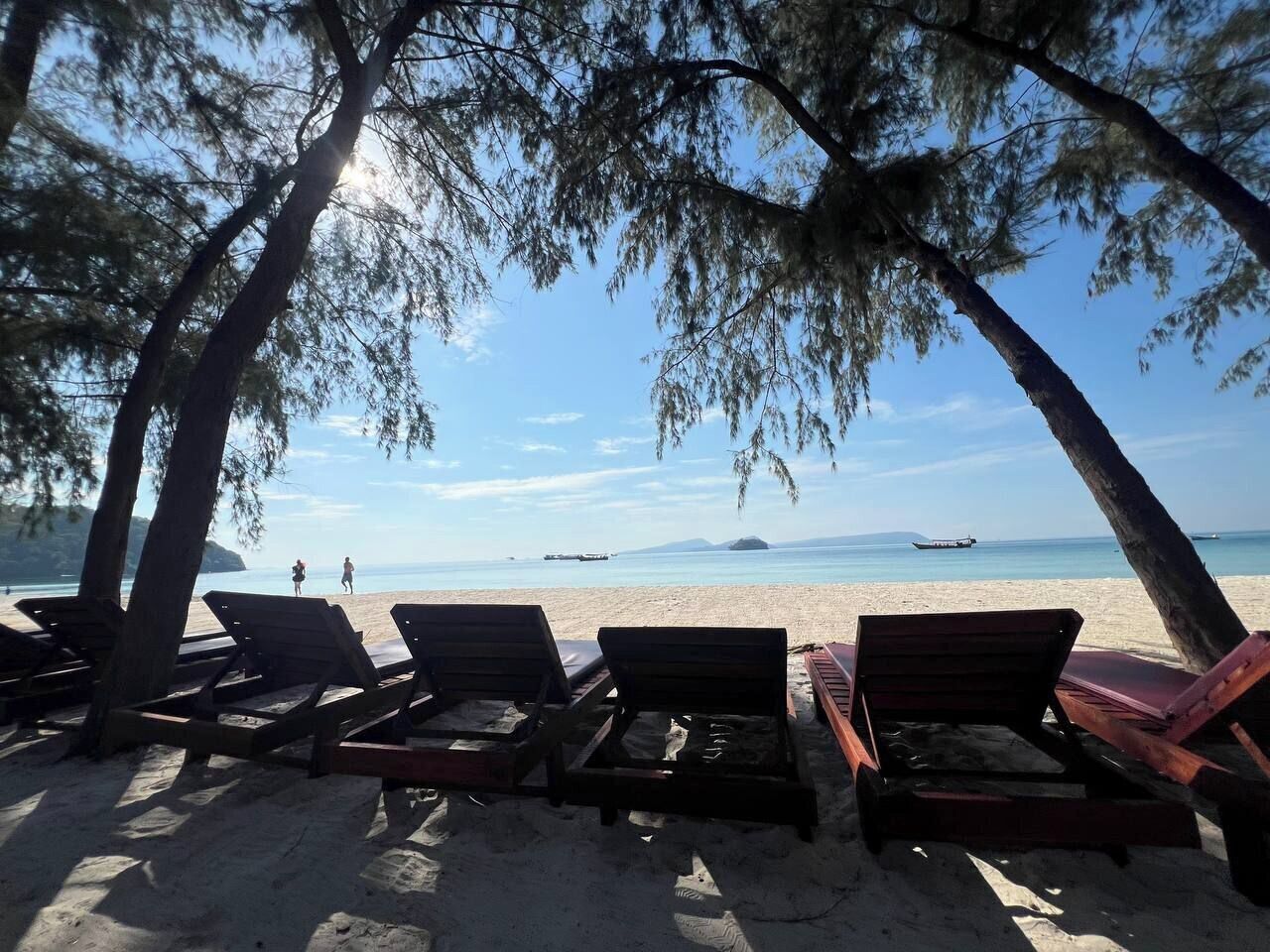 En strand med träsolstolar under träd, en klar himmel och ett lugnt hav.