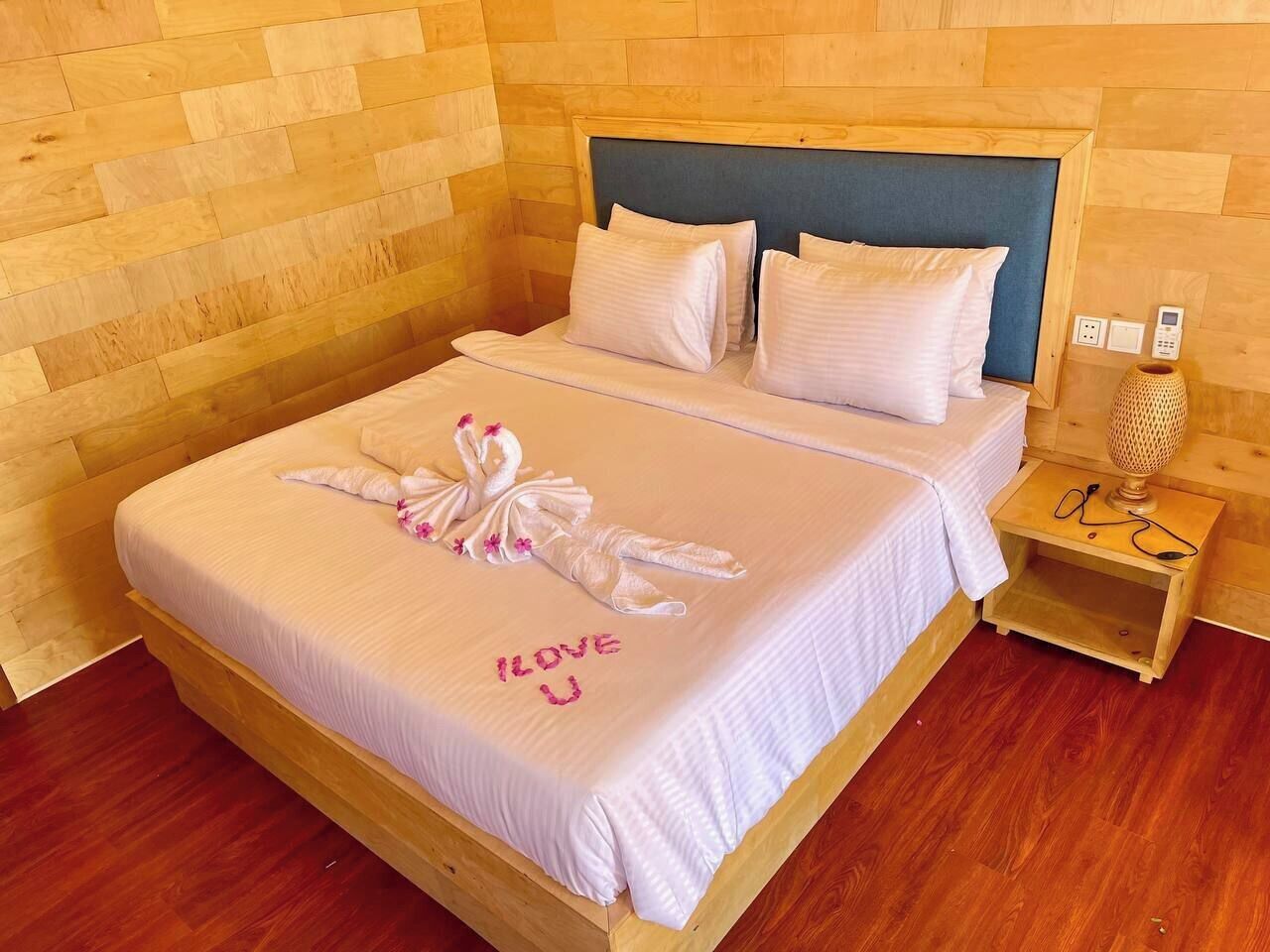 A neatly made bed with a towel and "I LOVE U" written on the bedspread, a bedside table with a lamp, and a wooden headboard.