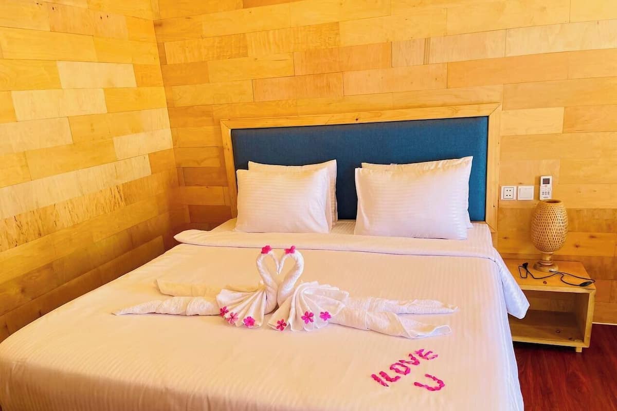 Quarto de hotel com parede de madeira, cama com roupa de cama branca, duas almofadas, mesa de cabeceira com abajur e arte de toalha na cama.
