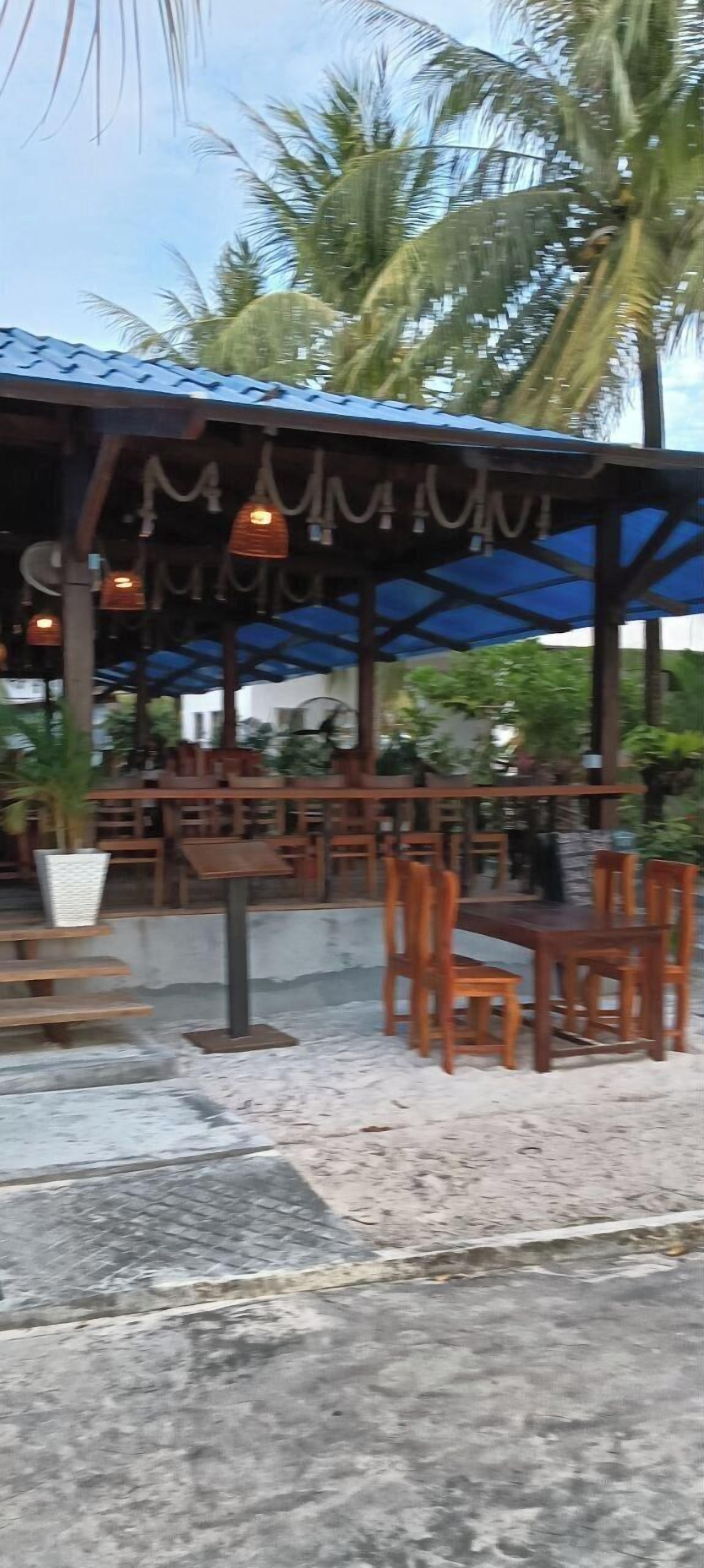 Een terras met houten tafels en stoelen, een overdeeld gedeelte met hangende lampen, en een achtergrond van palmbomen.