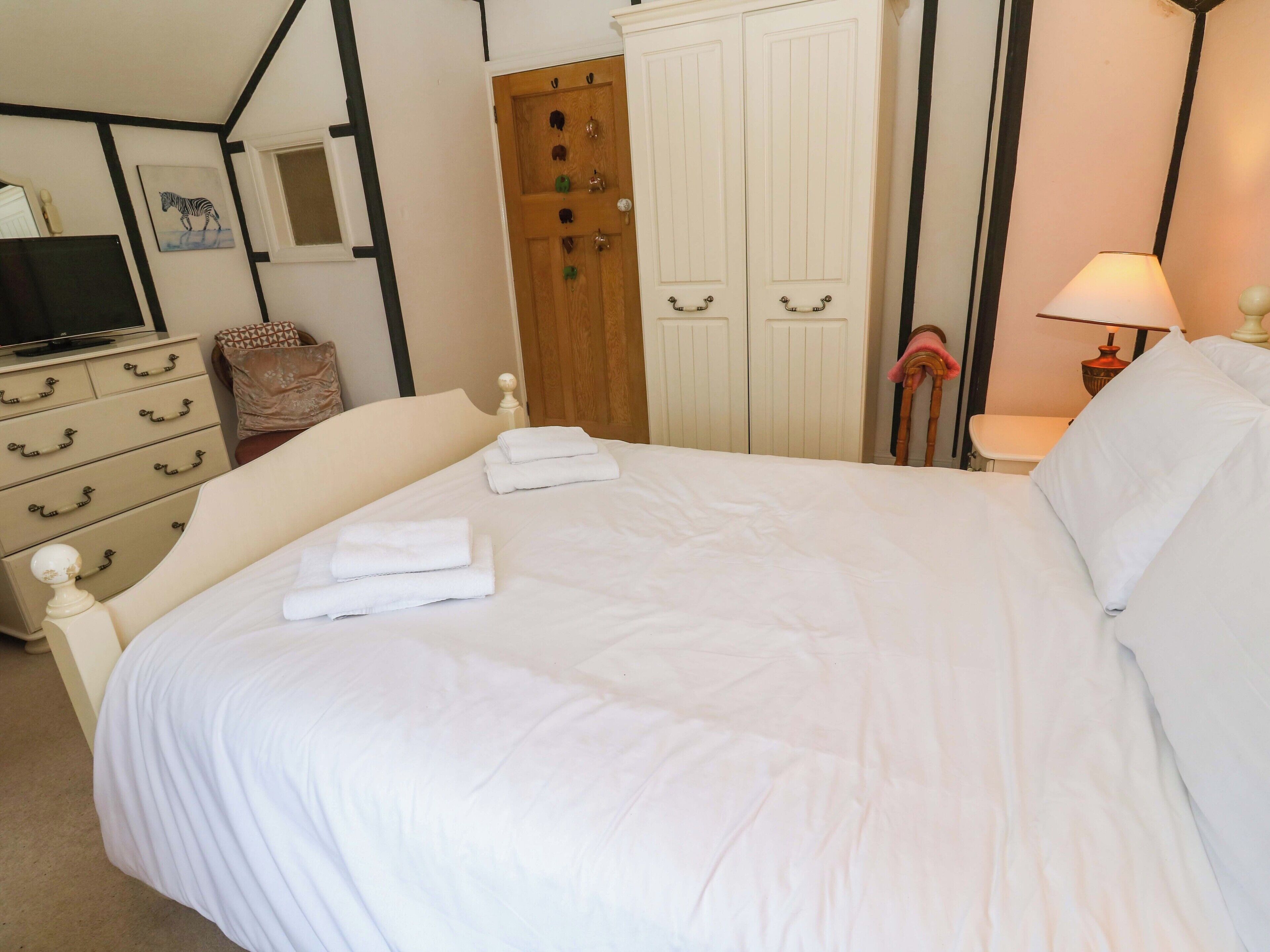 A neatly made bed with white linens and towels, a television on a dresser, a wooden wardrobe, and a lamp on a bedside table.