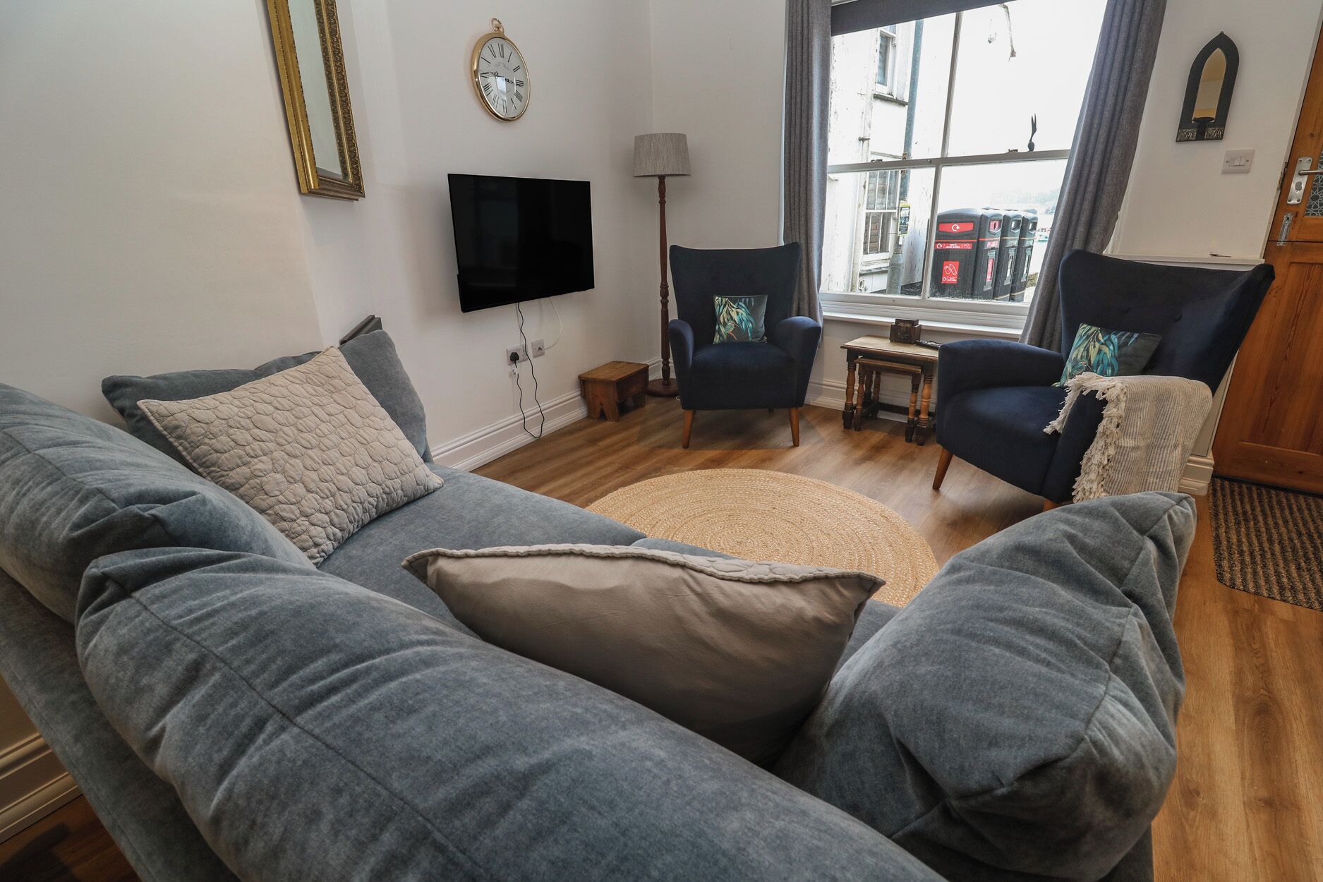 A living room with a grey sofa, two armchairs, a TV, a clock, and a window with a view of the street.