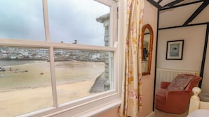 A room with a large window overlooking a beach, a red armchair, and a framed picture on the wall. - Pelican Cottage (St Ives)