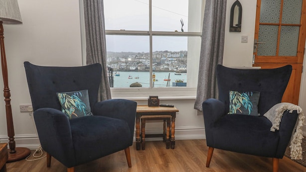 A room with two dark blue armchairs with patterned cushions, a small wooden table, a lamp, and a view of a harbor through a large window. - Pelican Cottage (St Ives)