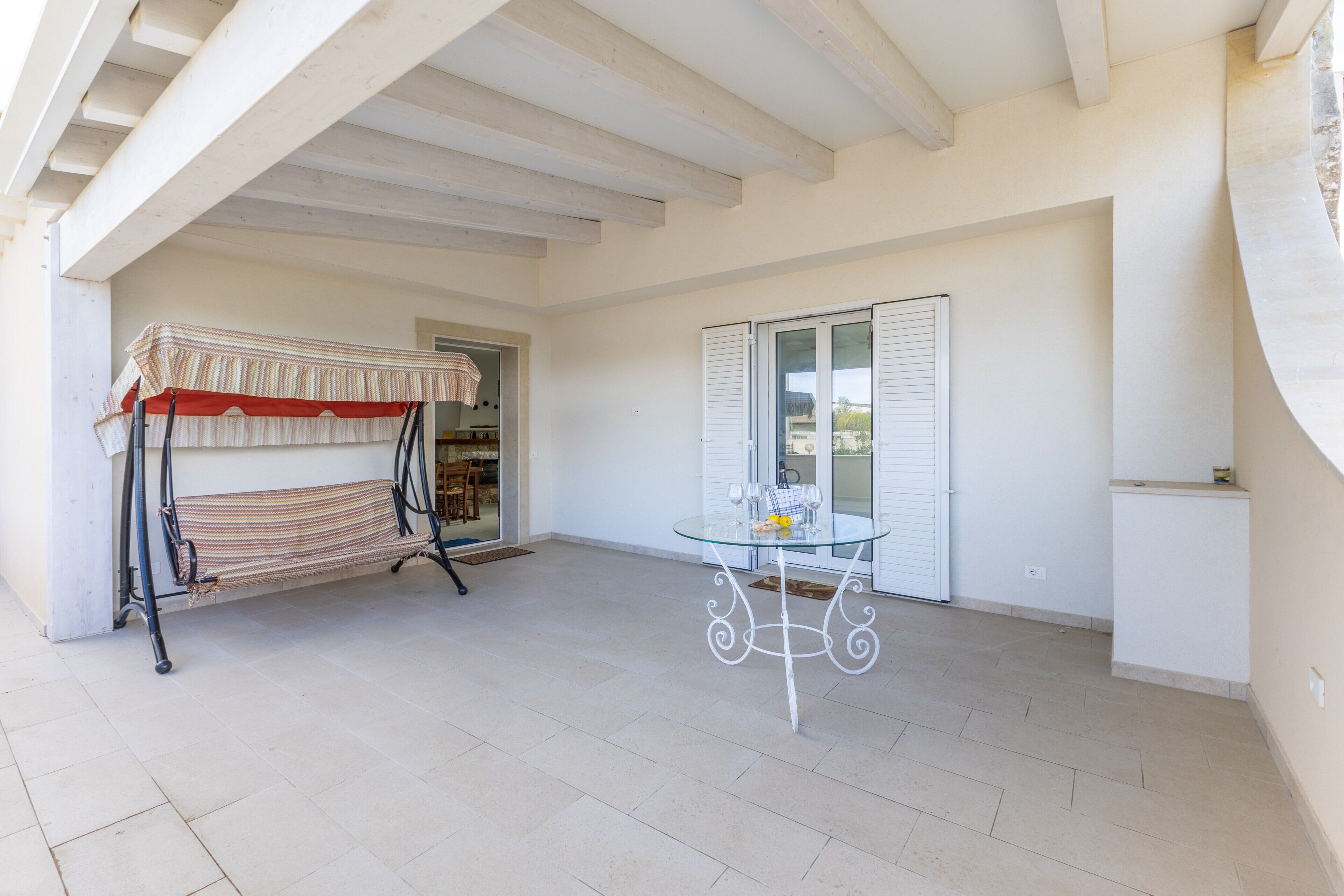 An open patio with a hammock, a glass table, and a door leading to a room.