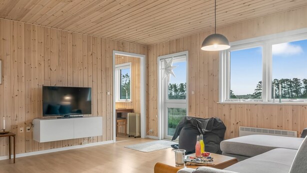 Un salon moderne avec des murs en bois, un téléviseur à écran plat posé sur un meuble blanc, un canapé gris et une grande fenêtre donnant sur la forêt.