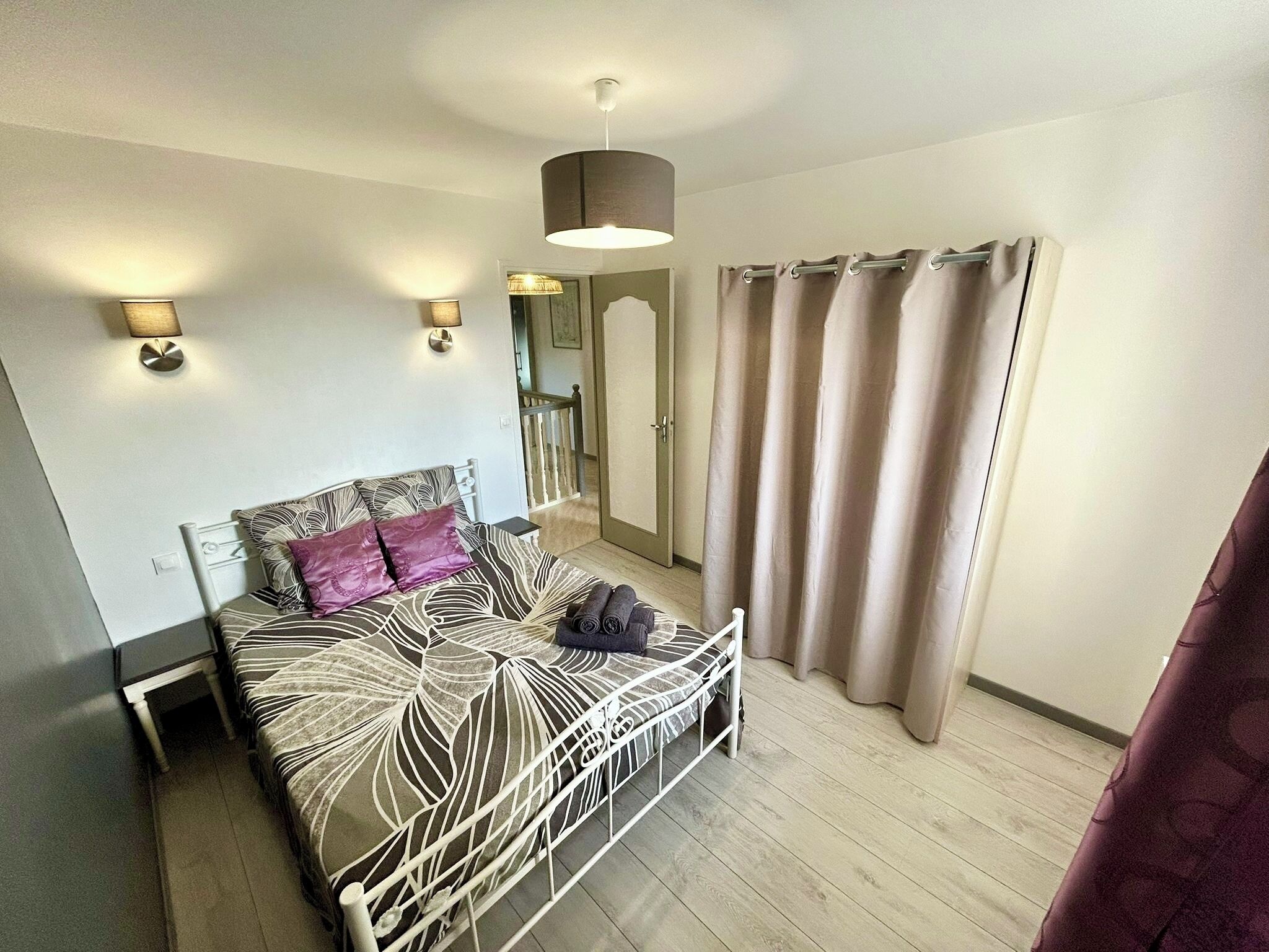Un dormitorio con cama, lámparas de noche, una lámpara colgante, una ventana con cortinas y una puerta que da a otra habitación.