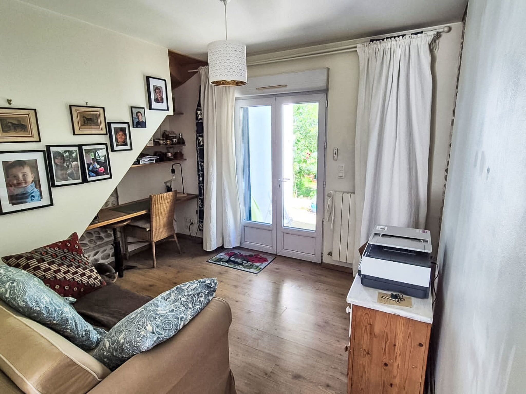 A cozy living room with a sofa, a desk, and a printer. There are framed pictures on the wall, and a door leading outside.