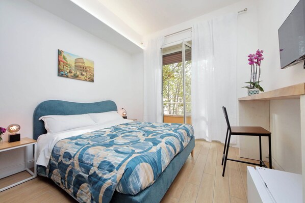 Une chambre à coucher avec un lit, un bureau, une chaise, un téléviseur et un tableau représentant un bâtiment.