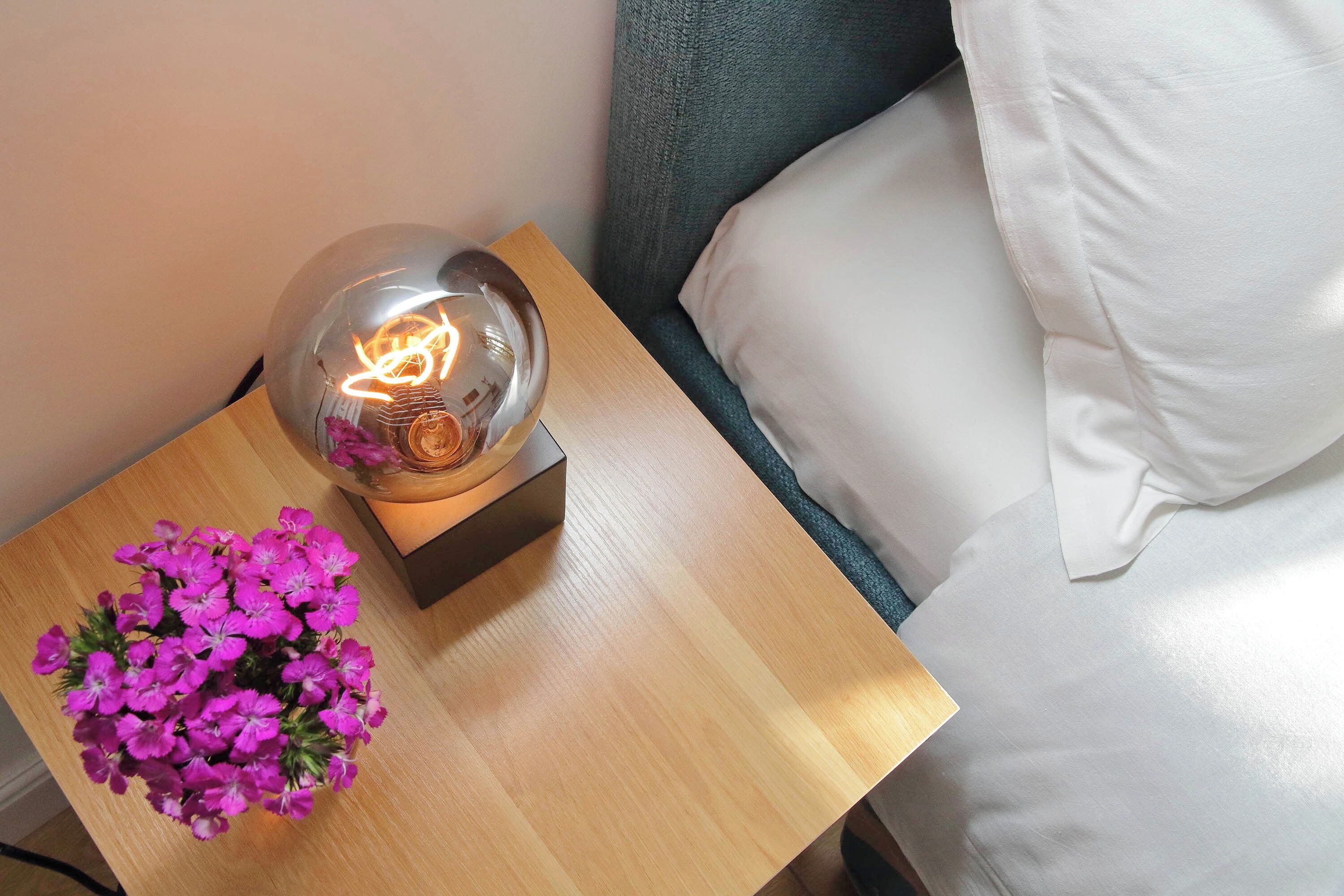 A wooden bedside table with a clear glass lamp containing a glowing light bulb and a small flower arrangement.