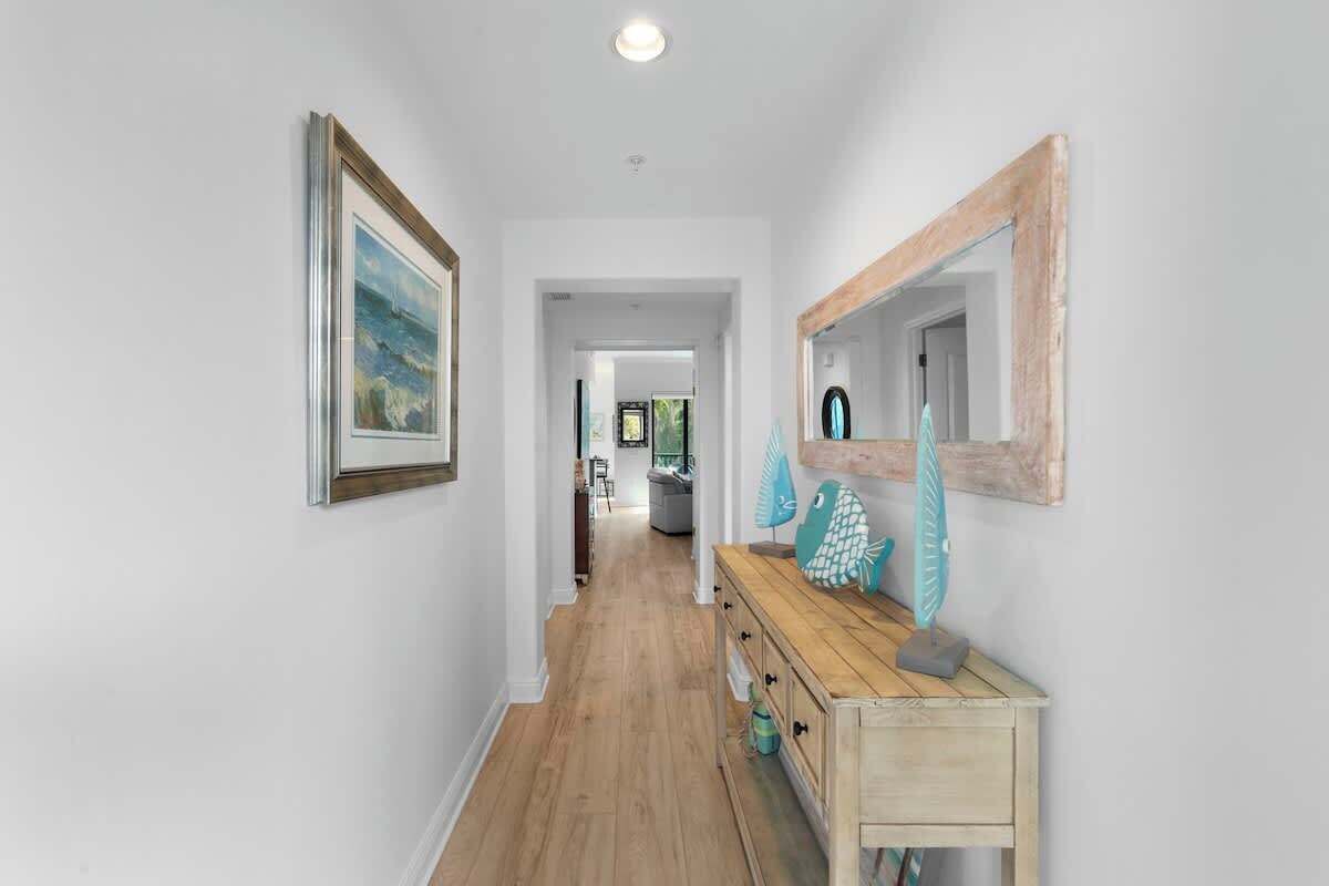 A hallway with a wooden floor, a wooden dresser with decorative items, a framed picture on the wall, and a mirror with a rustic frame.