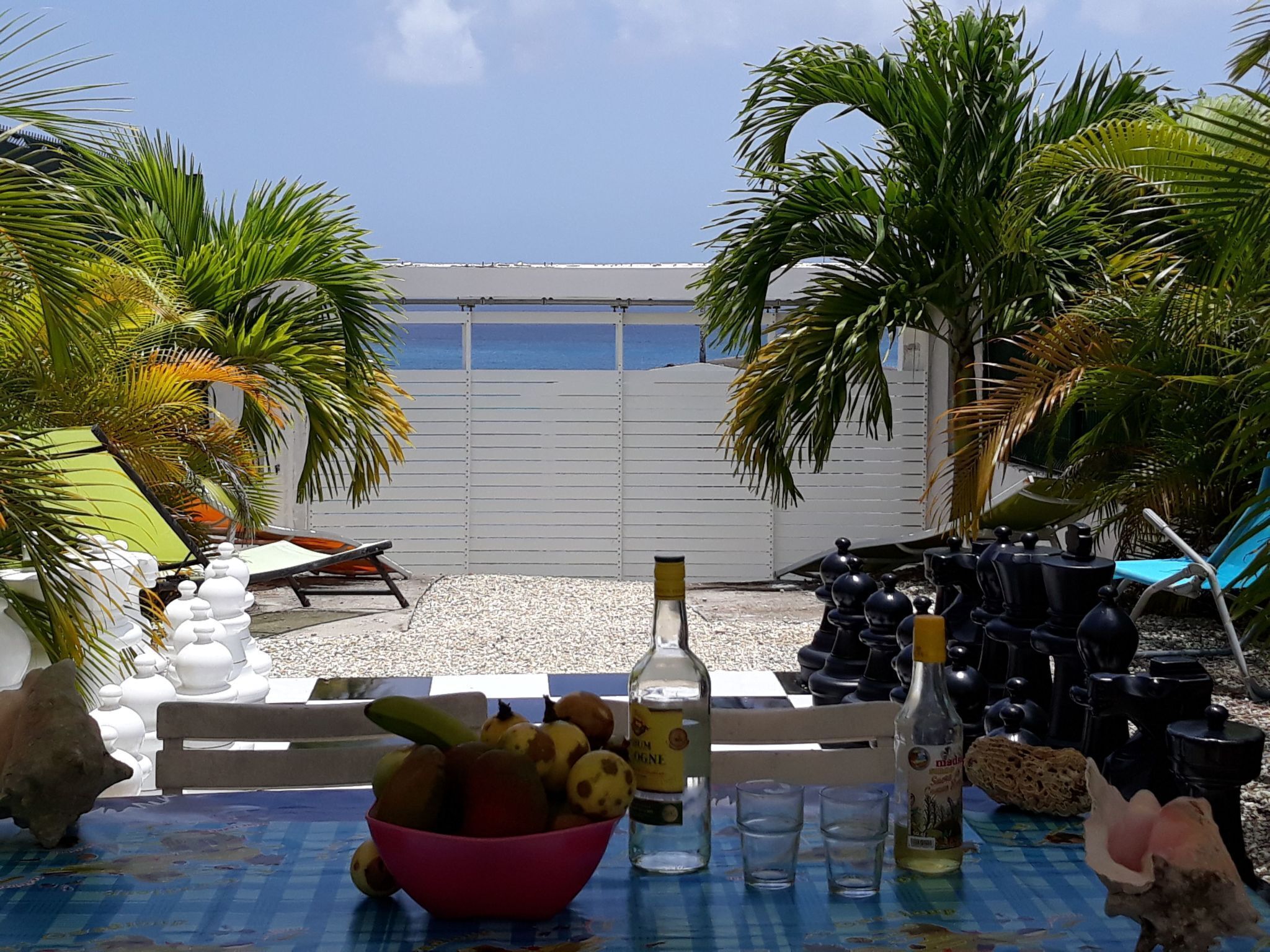 Ein Tisch mit einer Schale Früchte, Flaschen und Gläsern mit Blick auf einen Strand mit Palmen und einem weißen Zaun.