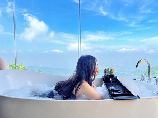 Eine Frau liegt in einer Badewanne, neben ihr ein Smartphone und ein Glas Wein, umgeben von Blasen. Durch große Fenster ist eine schöne Aussicht auf Berge und Himmel zu sehen.