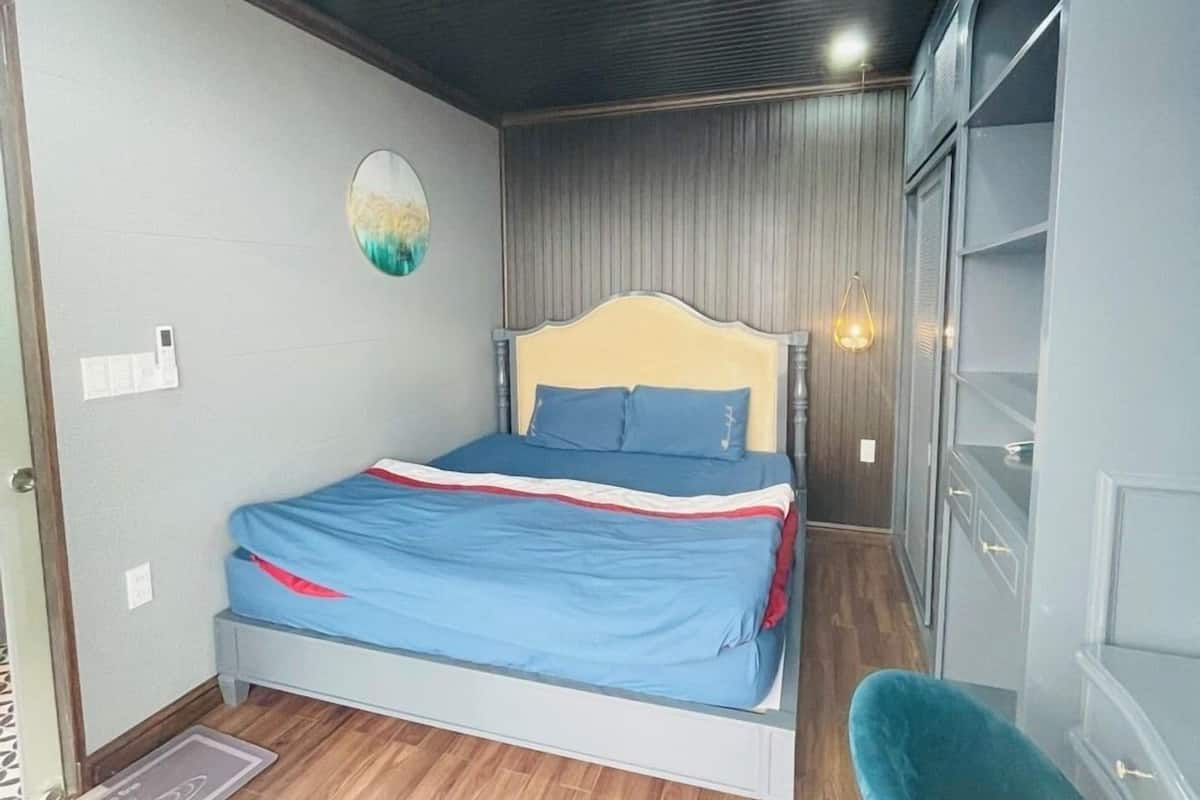 Un dormitorio con cama, una silla azul, un armario y un espejo redondo pequeño en la pared.