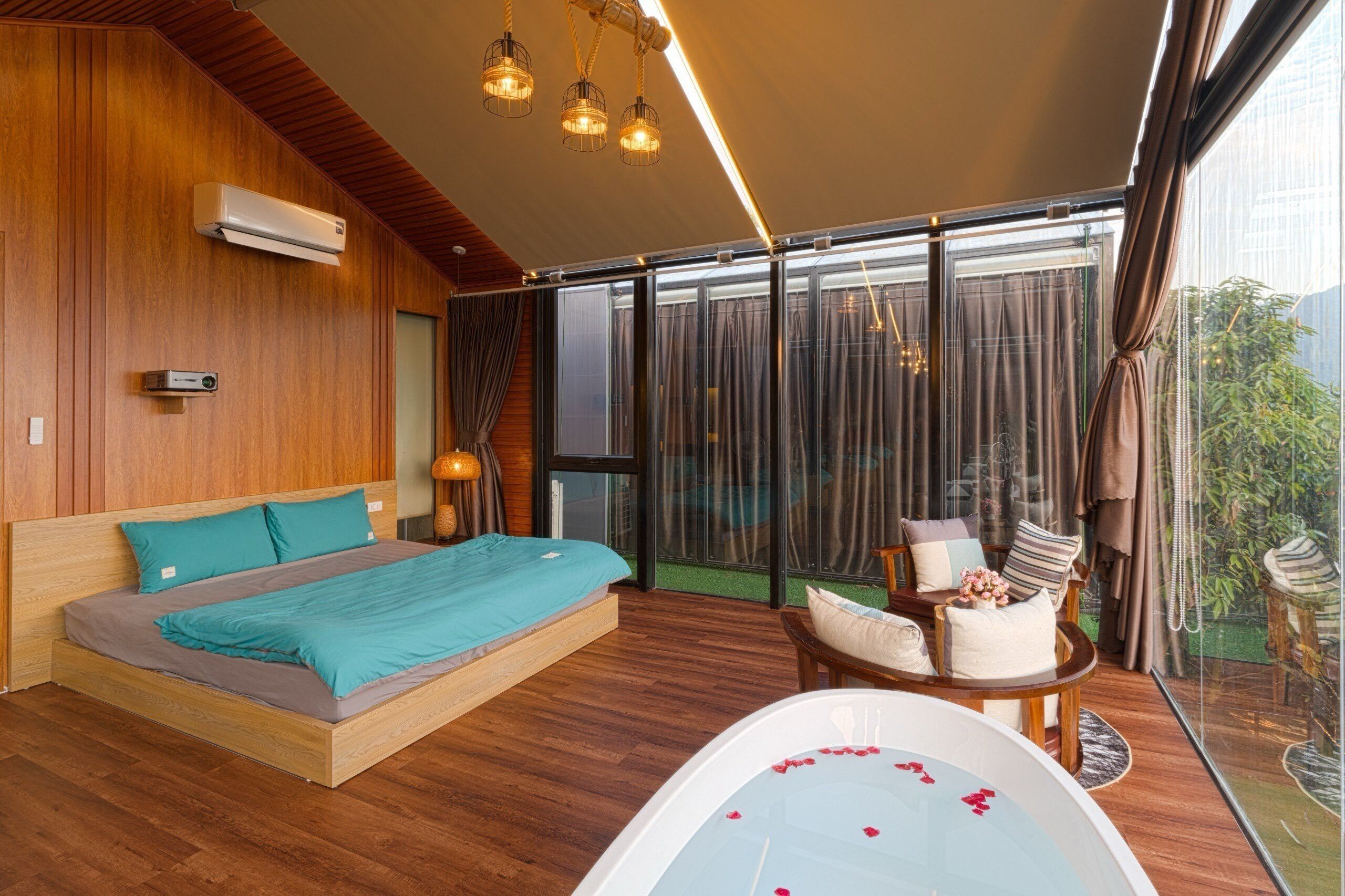 A modern bedroom with a large bed, a sitting area with a table and chairs, a bathtub with rose petals, and a view of greenery through large windows.