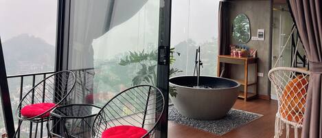 Un balcón con cerramiento de vidrio, una silla de metal negro con cojín rojo, una planta grande en maceta y una silla colgante con cojín de macramé.