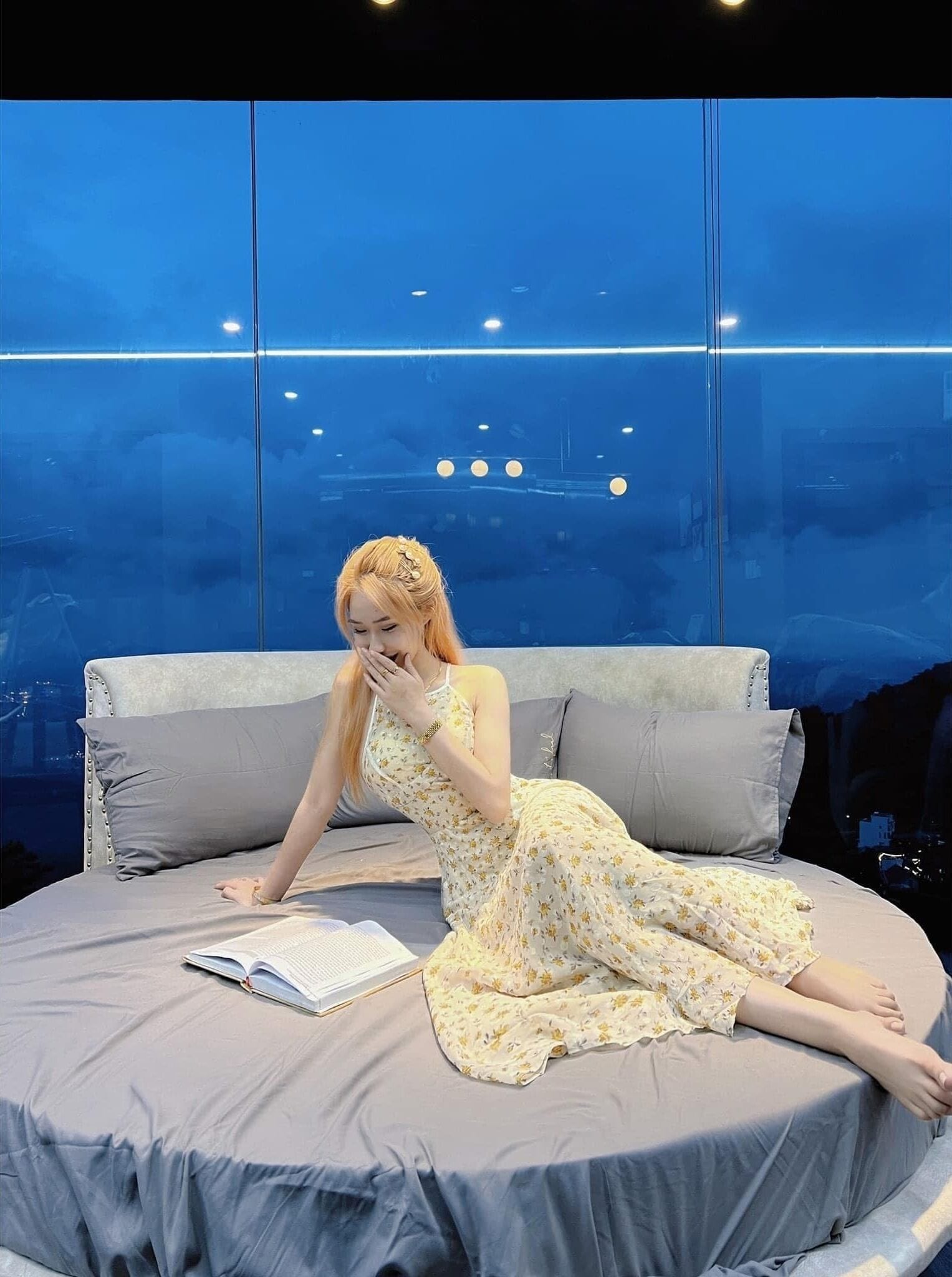 A woman sitting on a bed with a book, in a room with a large window and a view of the sky.