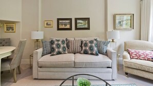 A living room with a beige sofa, a dining table, and chairs. There are framed pictures on the wall and a lamp on a side table. - Studio Flat With Garden Access, Holland Park (London)