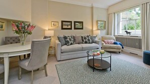 A living room with a sofa, a coffee table, a dining table, and a window with a view. - Studio Flat With Garden Access, Holland Park (London)