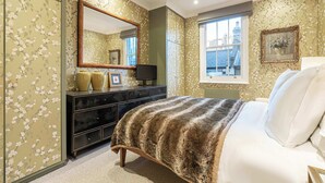 Una camera da letto con carta da parati a motivi floreali, un letto grande con coperta fantasia, un comò con specchio e una finestra vista edifici.