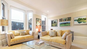 Un soggiorno luminoso con due divani beige, un tavolino in vetro e una libreria. Grandi finestre offrono luce naturale e vista sull'esterno.
