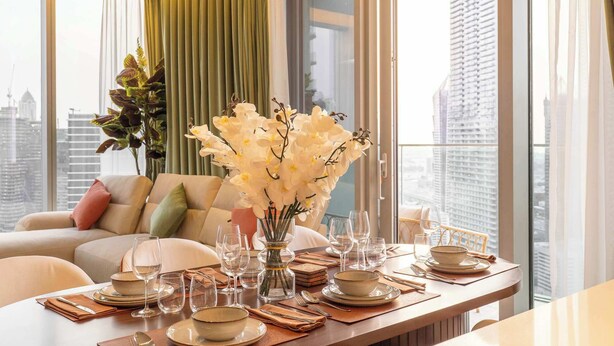 Comedor con una mesa dispuesta para cuatro, un jarrón de flores blancas y vistas de la ciudad a través de amplios ventanales.