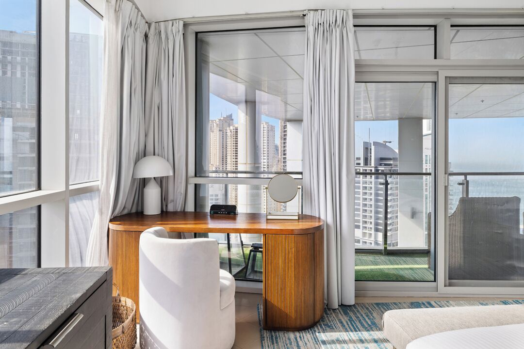 Quarto de hotel moderno com uma grande janela, uma escrivaninha de madeira, uma cadeira branca, uma cama e vista para a paisagem urbana.