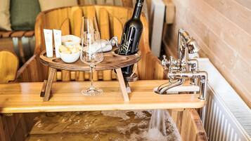 Una mesa de madera con una botella de vino, copas y un cuenco de fruta en una habitación con paredes de madera y una bañera.