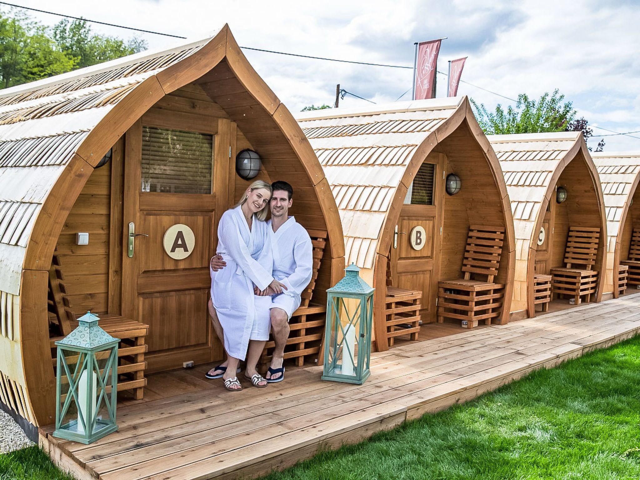 Una hilera de alojamientos tipo cápsula de madera, con puertas individuales marcadas con letras, una terraza de madera y faroles.