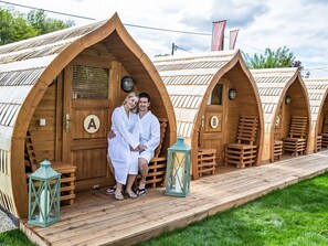Een rij houten accommodaties in de vorm van pods, elk met een deur voorzien van letters, een houten terras en lantaarns.