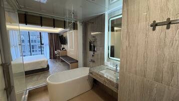 Een moderne hotelkamer met een groot bad, een wastafel met spiegel, een douche en een bureau met stoel, plus een bed.