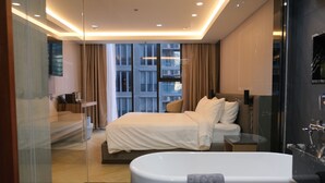 A modern hotel room with a large bed, a glass-enclosed shower area, and a view of the cityscape.