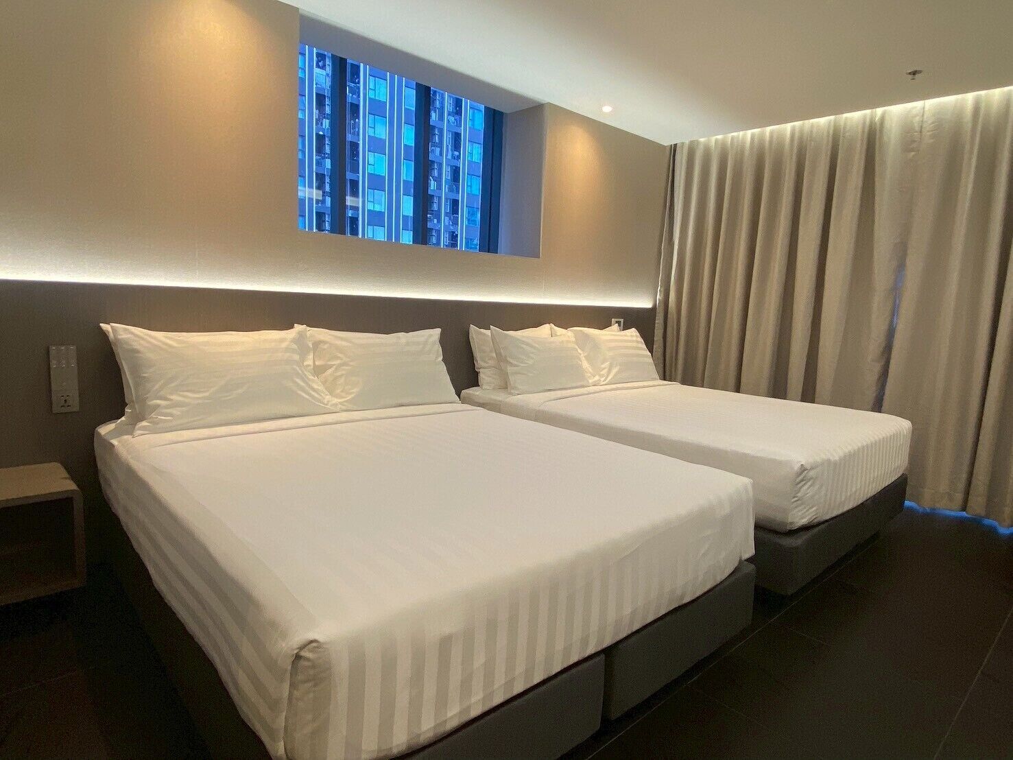 Habitación de hotel con dos camas, un ventanal con vistas a edificios y una cortina a la derecha.