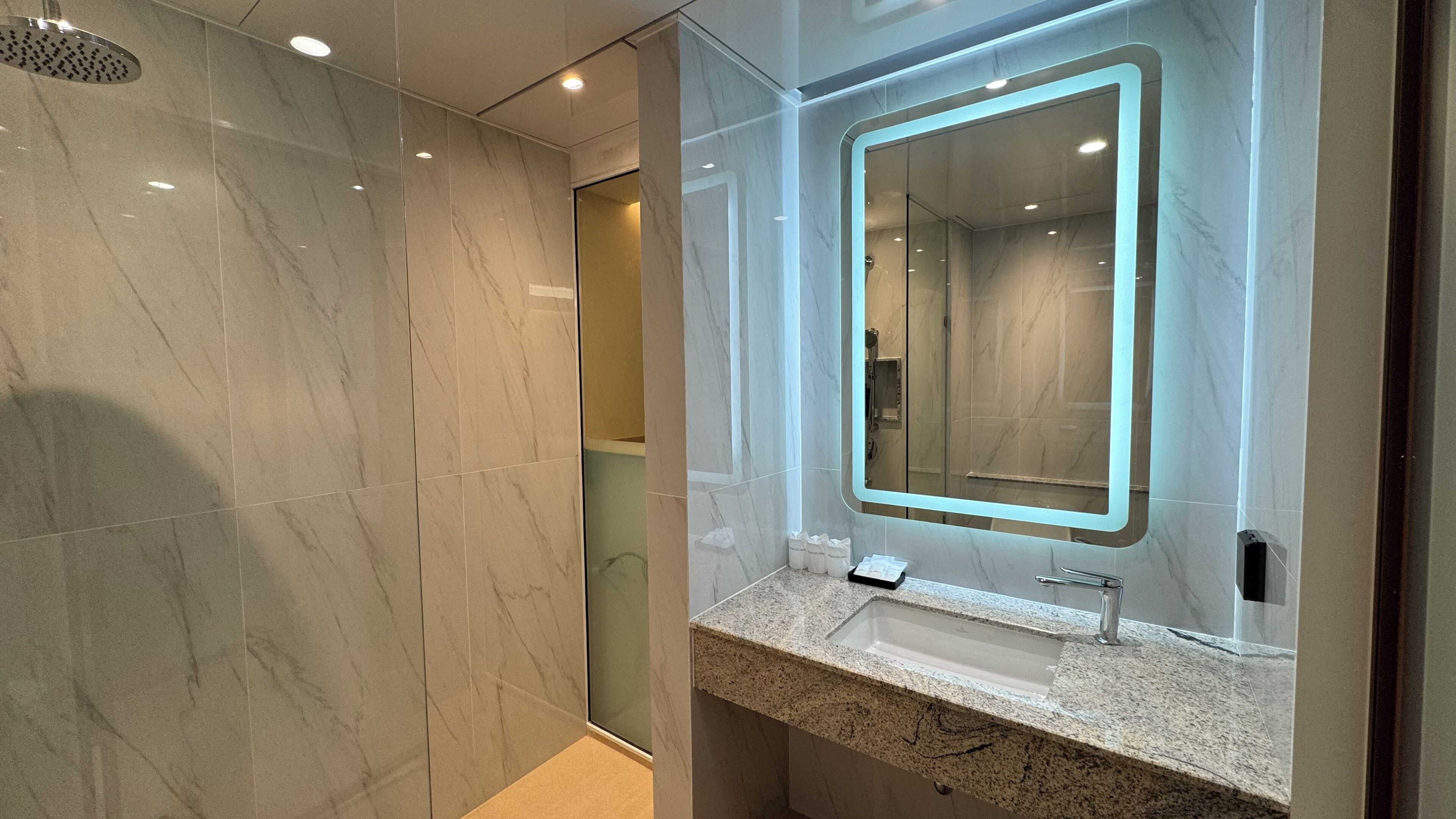 Un baño moderno con suelo de mármol, cabina de ducha de vidrio, lavamanos con espejo e iluminación empotrada.