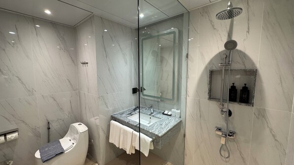 Un baño moderno con ducha envuelta en cristal, pared con azulejos de mármol, lavabo empotrado con encimera de mármol y un inodoro con dispensador de toallas de papel.