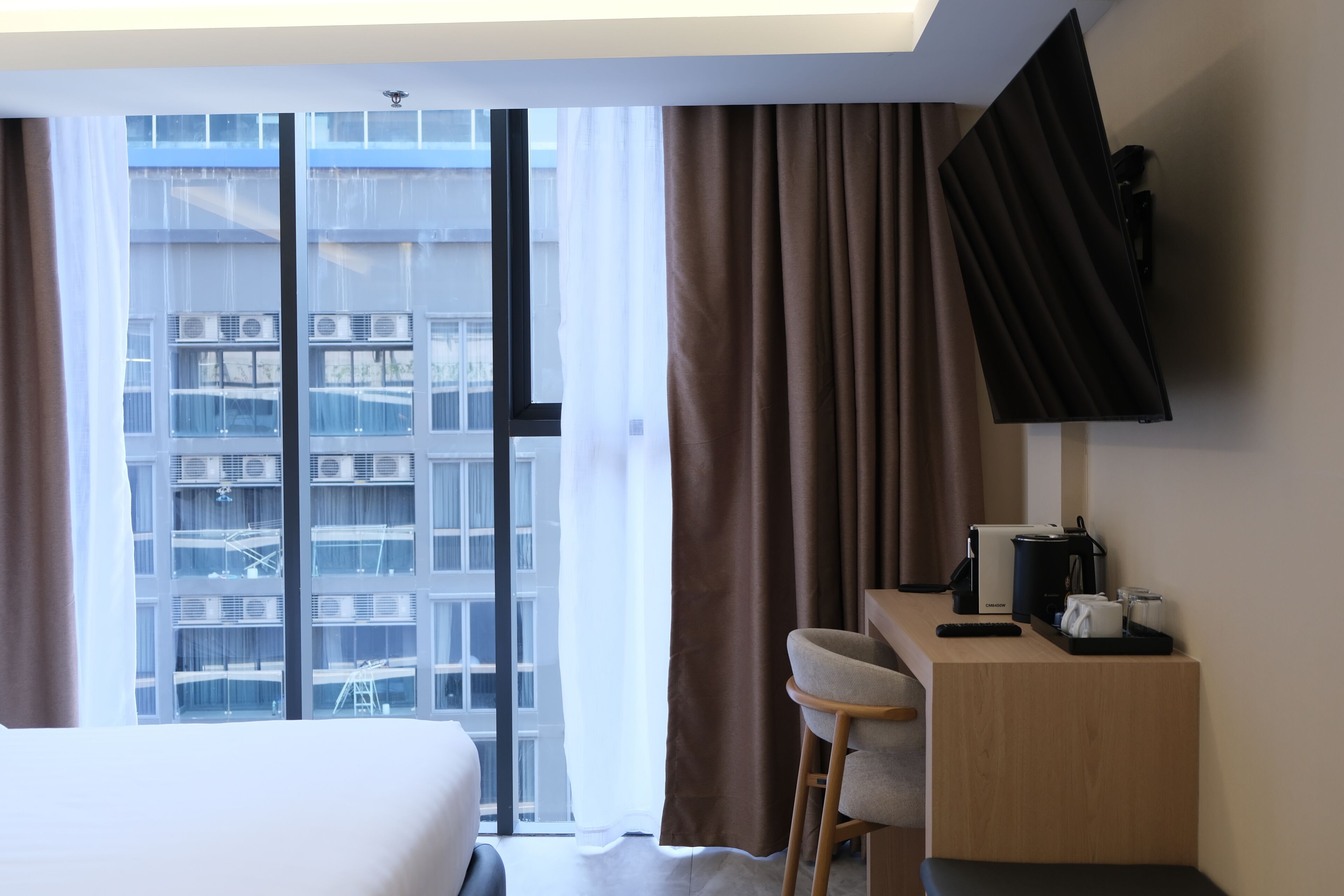 Ein Hotelzimmer mit einem Bett, einem Schreibtisch mit Stuhl, einem Flachbildfernseher und Blick auf ein Gebäude durch ein großes Fenster.