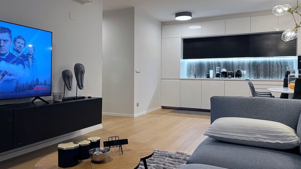 Een moderne woonkamer met een flatscreen televisie, een salontafel met boeken en een schaal, een grijze bank en een keukenhoek met een zwart aanrecht en witte kastjes.