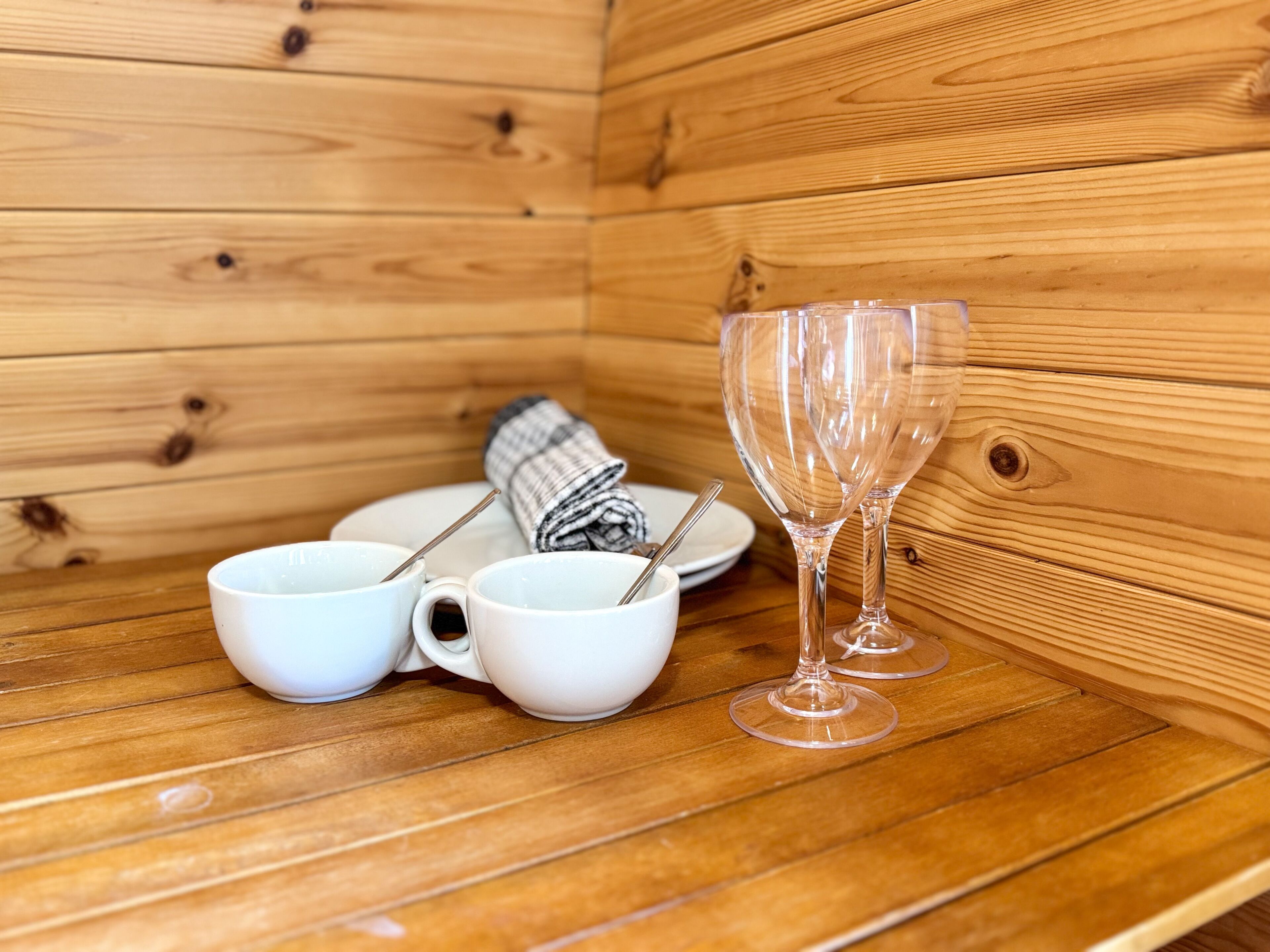 Interior de una cabaña de madera con un banco, dos tazas blancas con cucharas, una toalla enrollada y dos copas de cristal para vino.