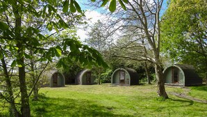 Une rangée de petites cabanes vertes, en forme de demi-cercle, se trouve dans une zone herbeuse bordée d’arbres.