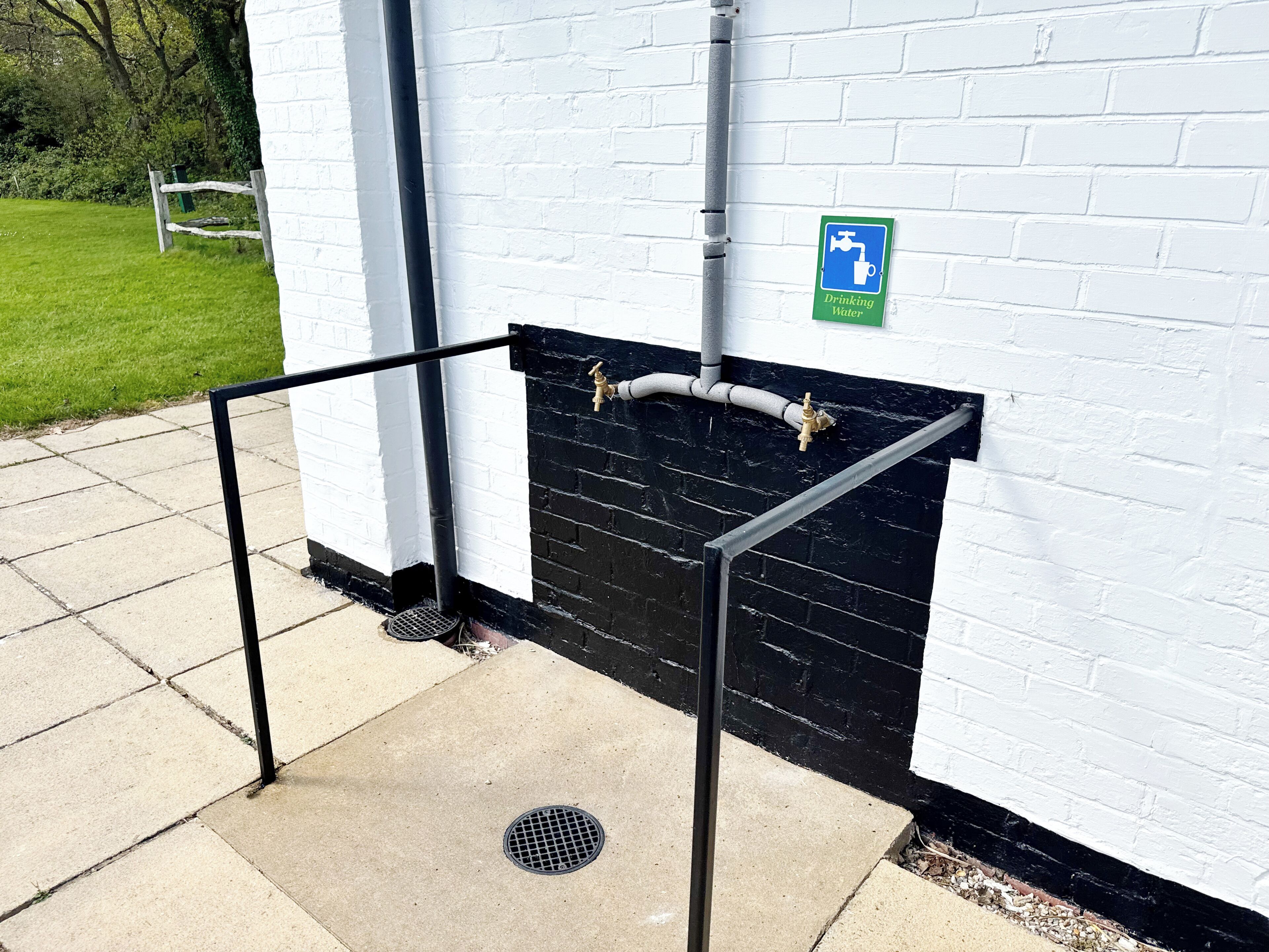 Eine öffentliche Trinkwasserstation mit Schild, Metallgeländer und einer schwarzen Backsteinmauer.