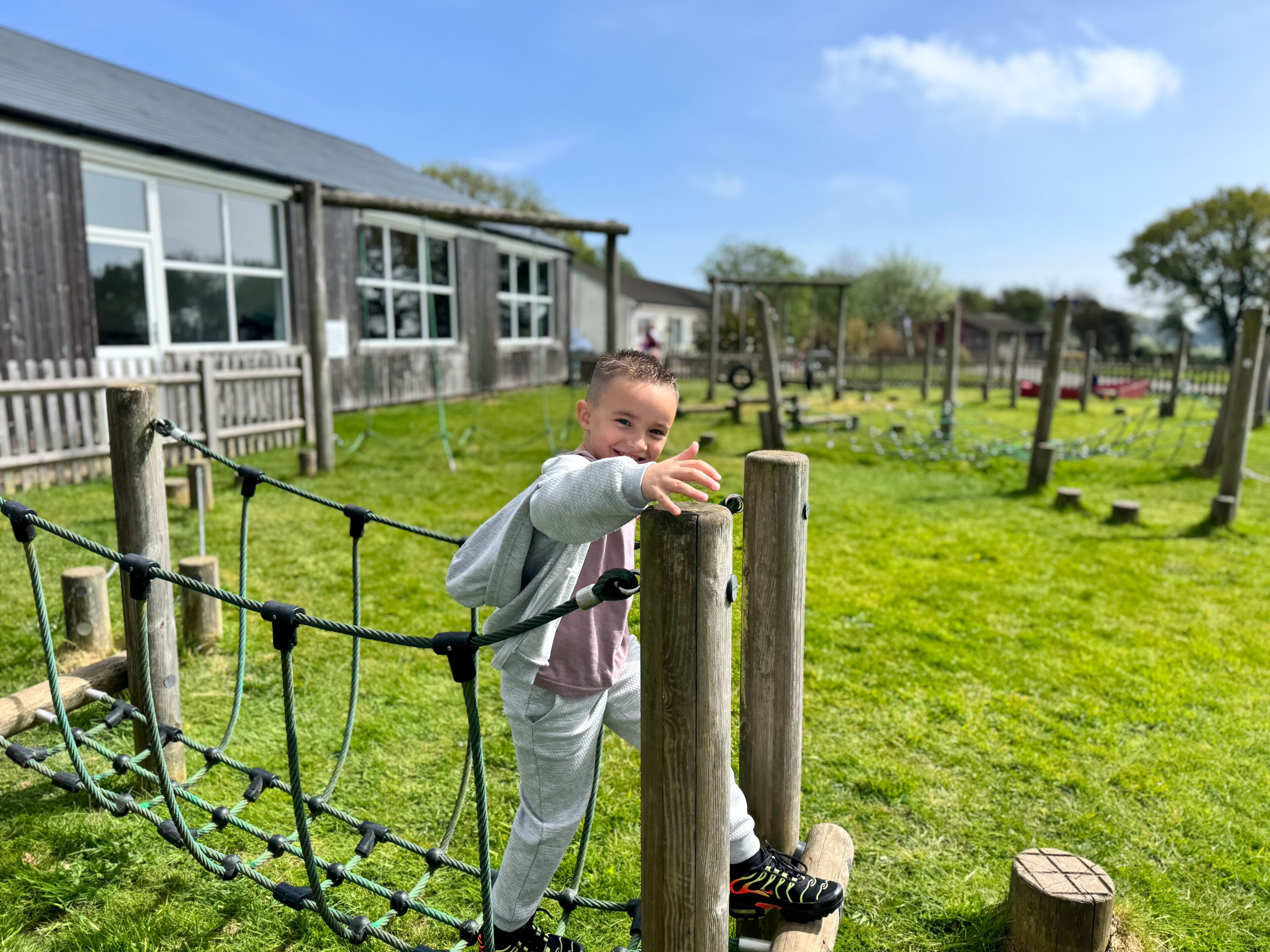 A child playing on a rope bridge in a grassy outdoor area with wooden structures and a building in the background.
