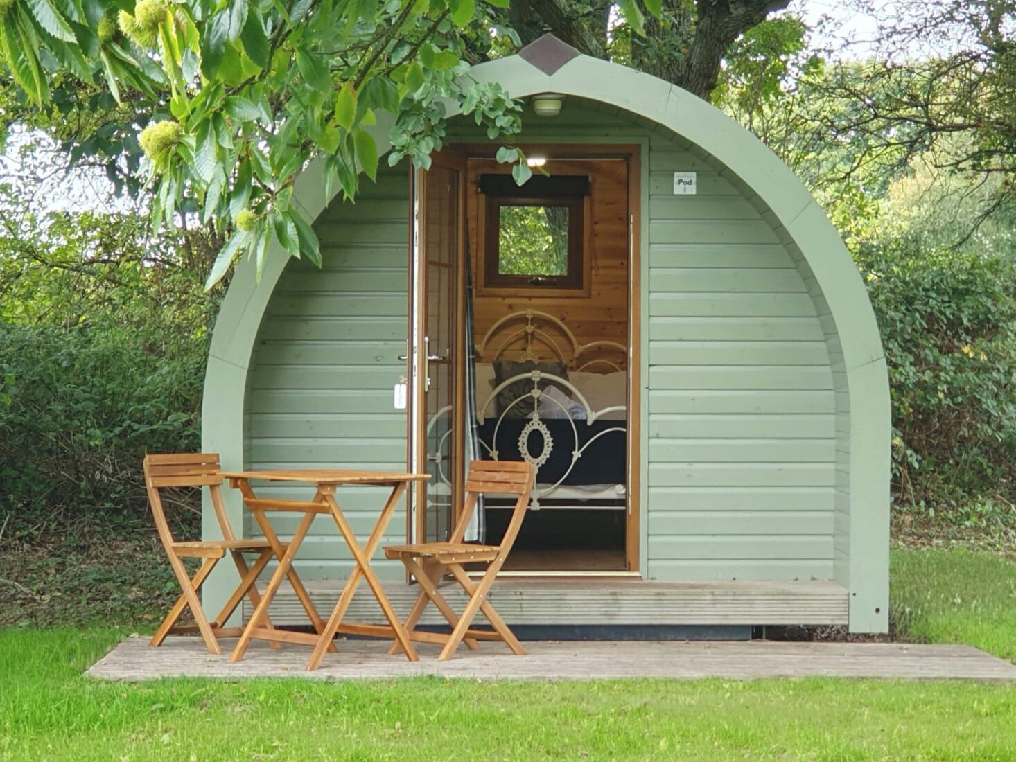 A small, green, semi-circular structure with a door, a bed inside, and two wooden chairs outside.